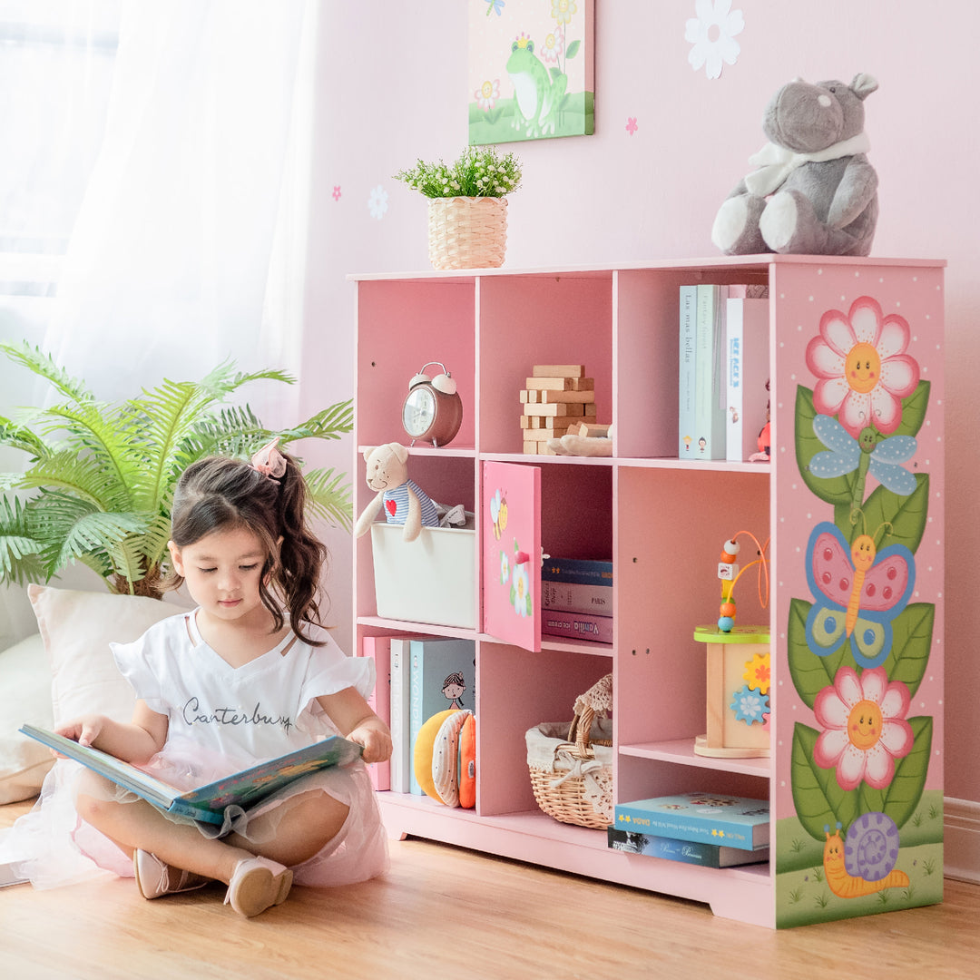 A girl sitting on the floor reading a book from her Fantasy Fields Kids Painted Wooden Magic Garden Adjustable Cube Bookshelf, Pink.