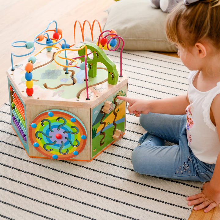 A child playing with a Teamson Kids Preschool Play Lab 7-in-1 Large Wooden Activity Station featuring durable construction, on a striped rug.