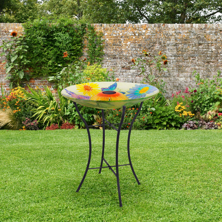 A colorful floral-patterned tabletop on a metal stand serves as an attractive Teamson Home Outdoor 18" Hand painted Butterfly Fusion Glass Solar Bird Bath in a garden, blending impeccably with outdoor furniture.