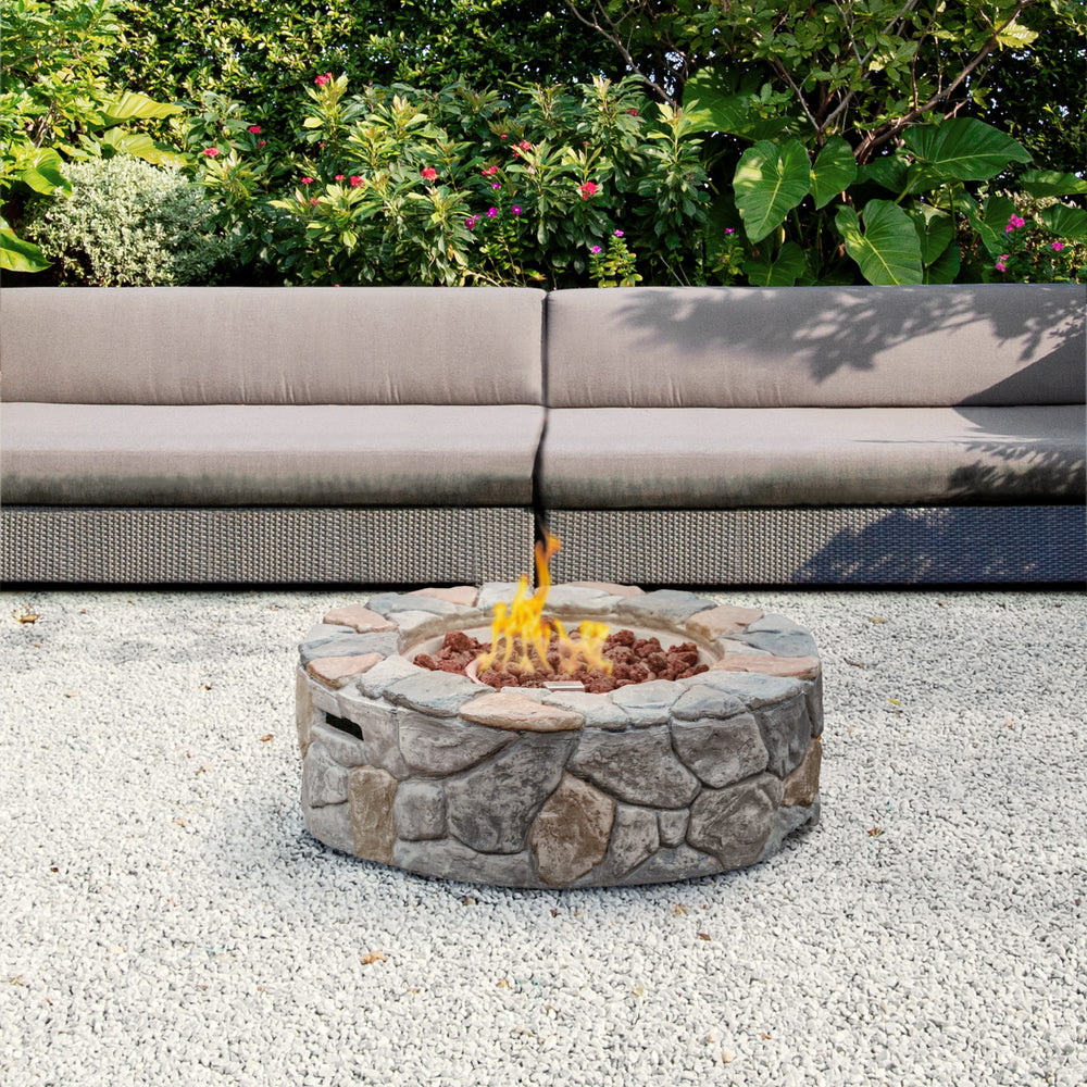 A Teamson Home 28" Outdoor Round Stone Propane Gas Fire Pit with flames alight, surrounded by outdoor seating in a garden setting.