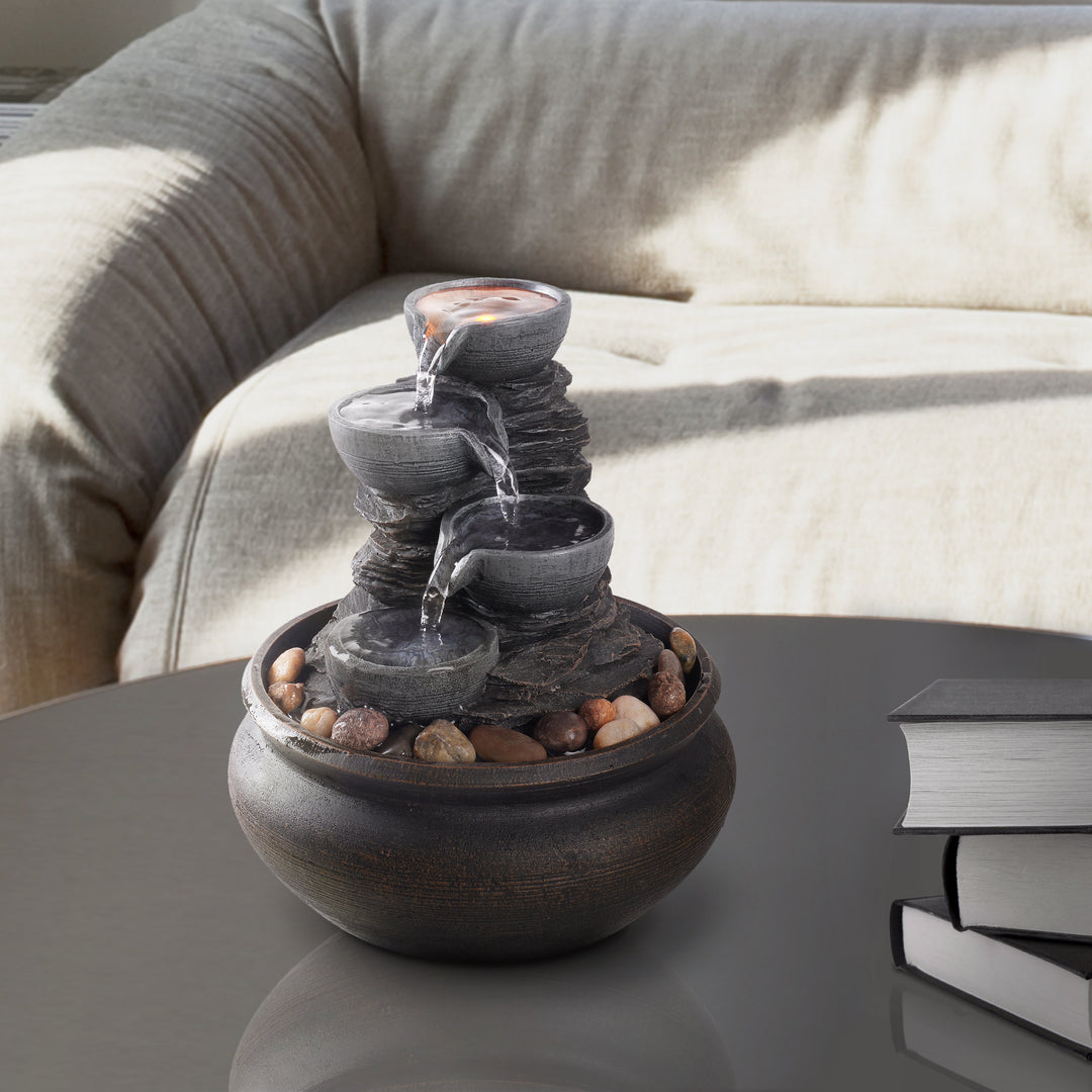 Teamson Home Tabletop Water Fountain, Stone Gray, with four tiers and pebbles on the base and a LED light in the top tier, on a coffee table next to books