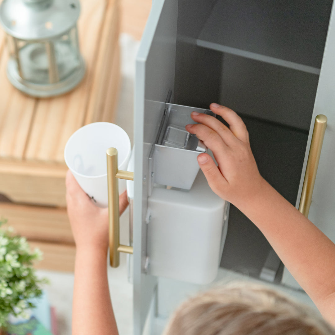 A child reaches for a water dispenser in a fridge while holding a white mug in their Teamson Kids Little Chef Charlotte Modern Play Kitchen, Silver Gray/Gold.