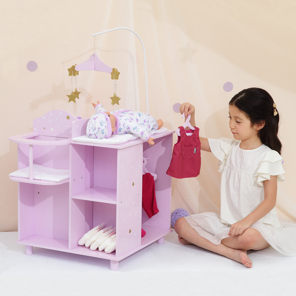 A little girl holding a red baby doll dress sitting next to a  baby doll changing station in purple with white and gold stars.