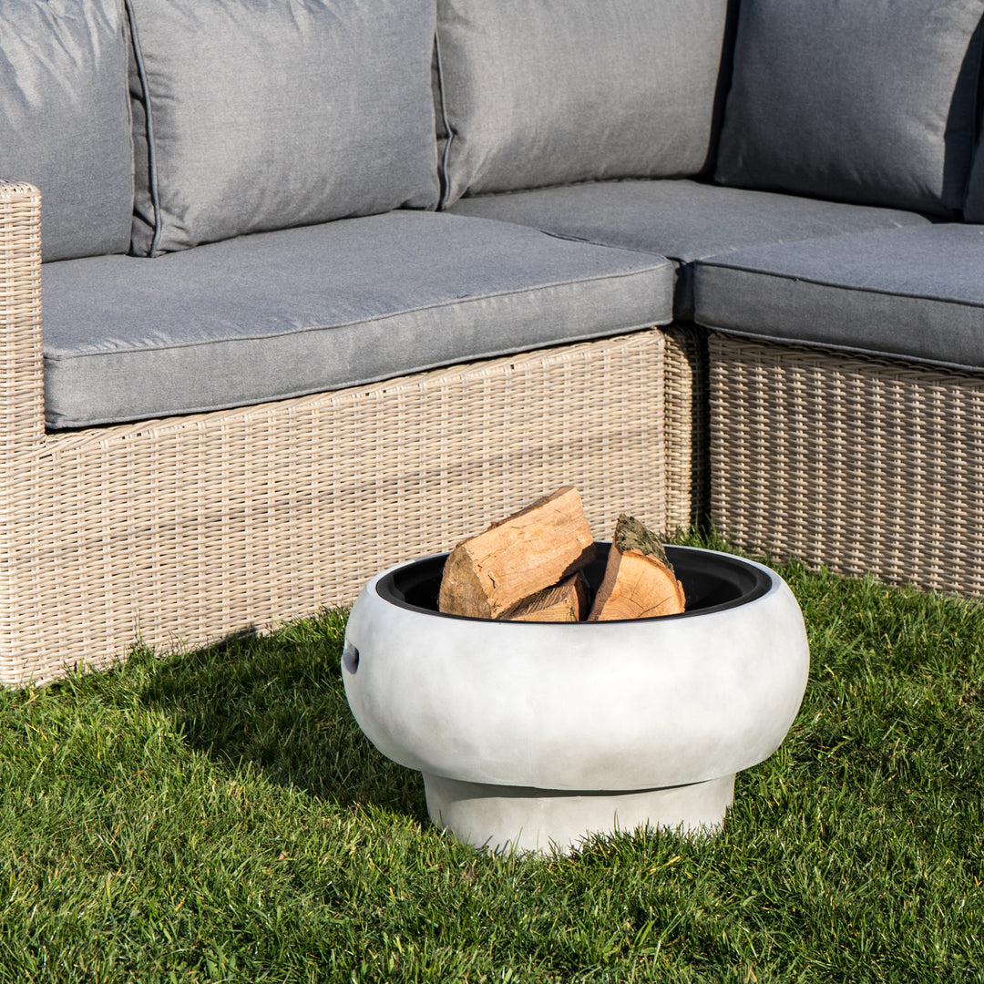A Teamson Home 21" Outdoor Round Stone Wood Burning Fire Pit with Faux Concrete Base, Gray with firewood in front of a patio sofa with cushions.