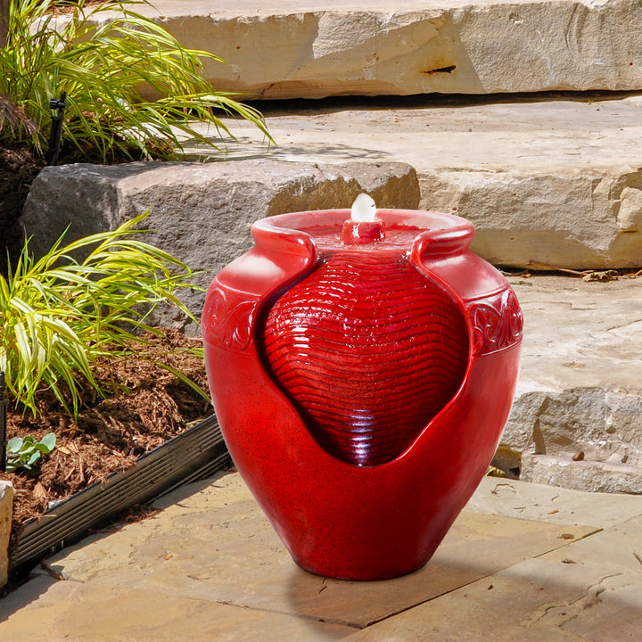 A Teamson Home Outdoor Glazed Pot Floor Fountain with LED Lights, Red, placed on a stone patio surrounded by greenery.