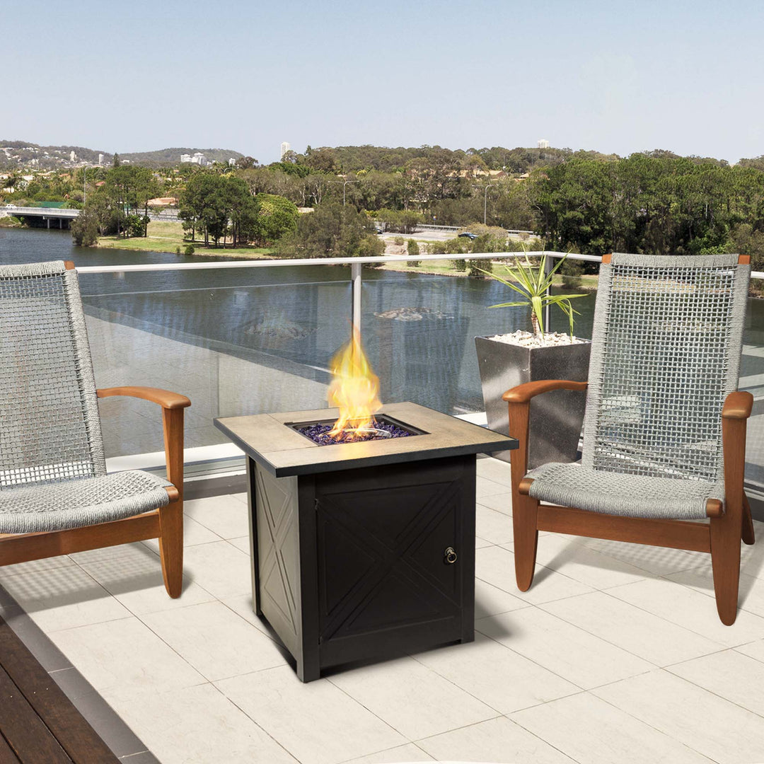 A Teamson Home Outdoor Square 30" Propane Gas Fire Pit with Steel Base, Black, with a pair of chairs on a deck overlooking a body of water