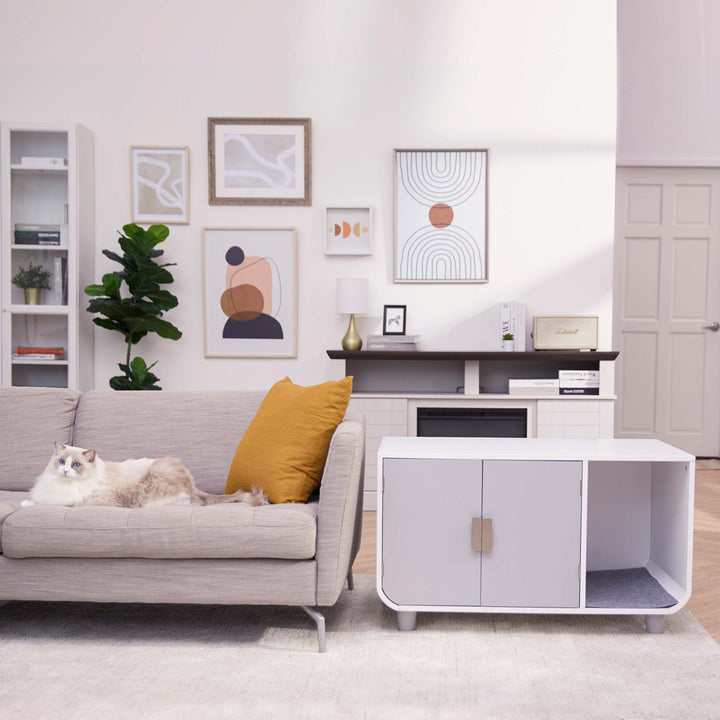 Teamson Pets Large Dyad Wooden Cat Litter Box Enclosure and Side Table, Alpine White/Gray next to a flax-colored sofa with a yellow pillow and white cat.
