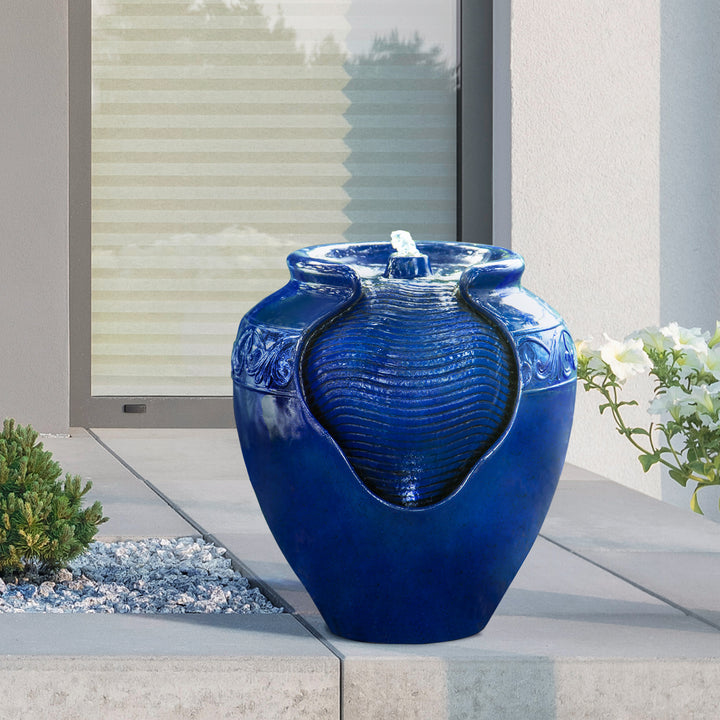 A Teamson Home Outdoor Glazed Pot Floor Fountain with LED Lights, Royal Blue placed on a concrete slab outdoors, with water flowing over its smooth surface, serving as charming garden decor.