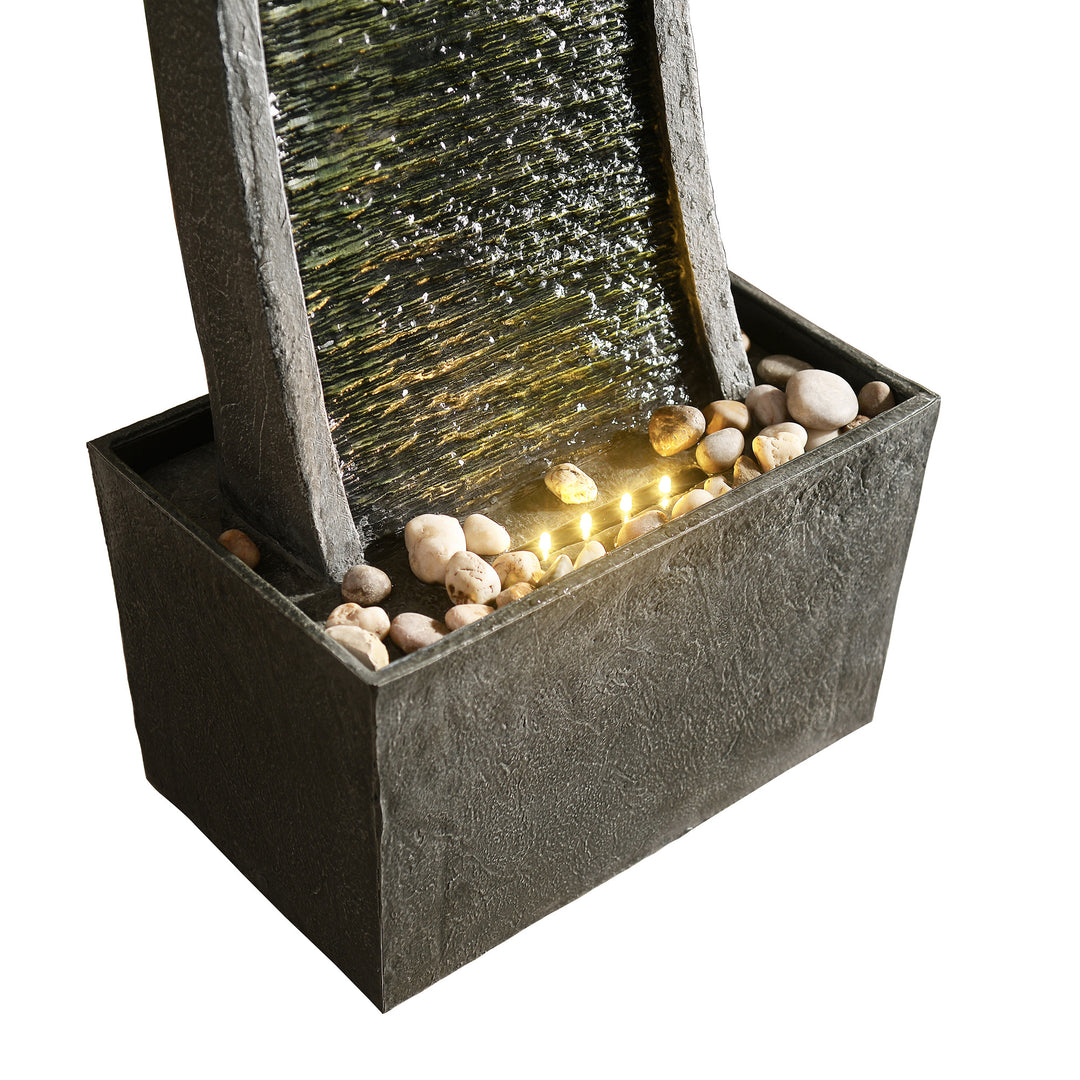 A Teamson Home Indoor/Outdoor Modern Curved Slate Stone-Look Tall Waterfall Fountain with LED Lights featuring a textured column with water cascading down, surrounded by smooth stones and illuminated by LED lights within a square basin.