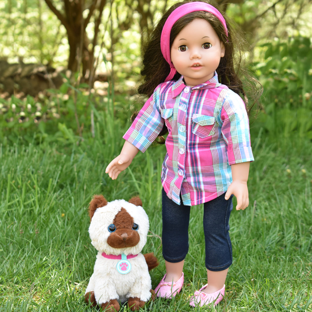 An 18" brunette doll with a plaid shirt and denim capri pants and pink shoes. She is standing on some grass with a siamese kitten.