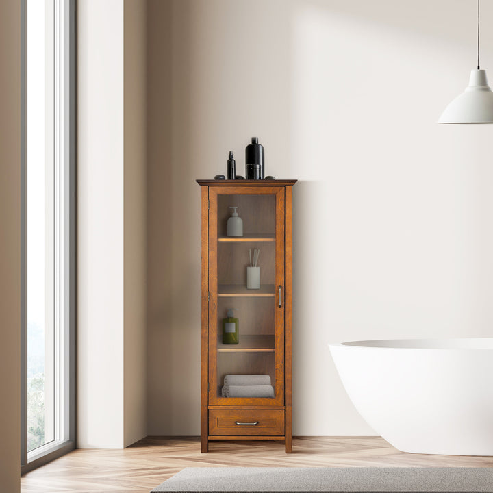 Teamson Home Avery Wooden Linen Tower Cabinet with Storage, Oiled Oak linen storage cabinet with toiletries next to a freestanding bathtub in a bright bathroom.