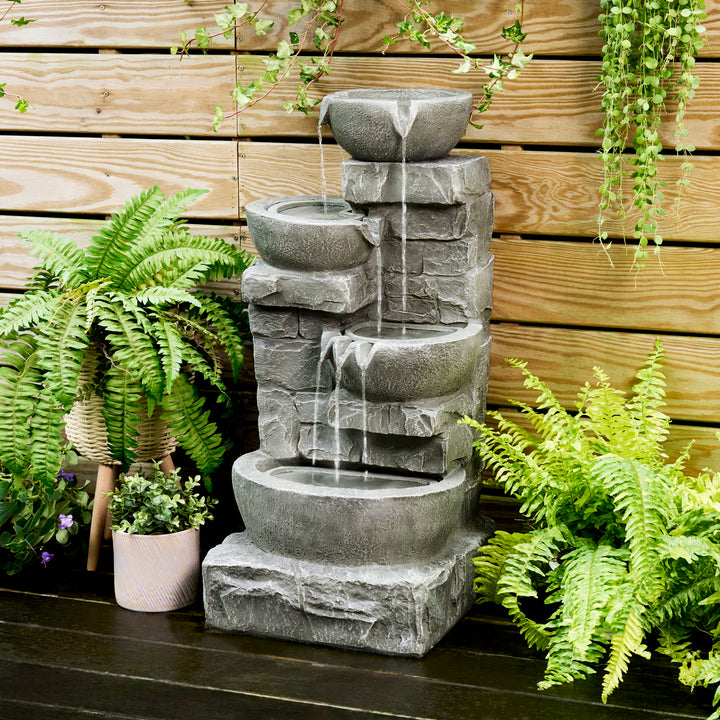 Teamson Home Outdoor Cascading Bowls & Stacked Stone water fountain set against a wood-planked wall amongst plants
