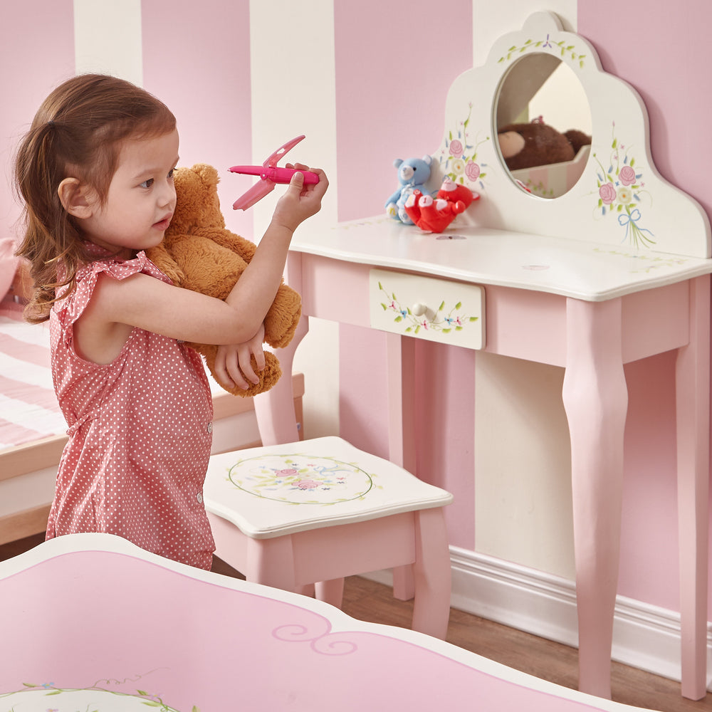 A little girl holding a teddy bear in a Fantasy Fields Kids Furniture Play Vanity Table and Stool-themed pink bedroom.