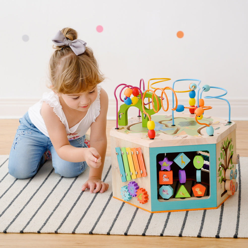 A young child playing with a Teamson Kids Preschool Play Lab 7-in-1 Large Wooden Activity Station on a striped rug.