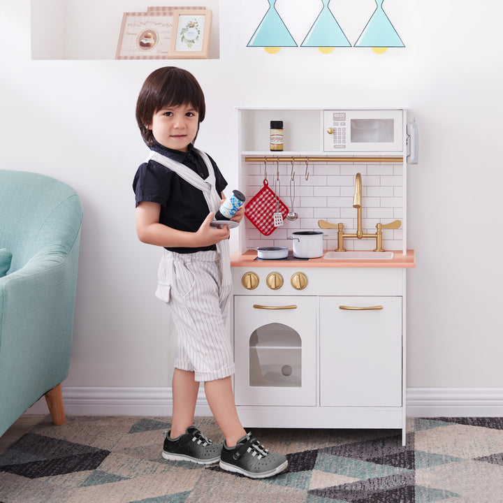 A child stands next to a Teamson Kids Little Chef Boston Classic Play Kitchen & Cookware, White, holding a bottle with a smile on their face.