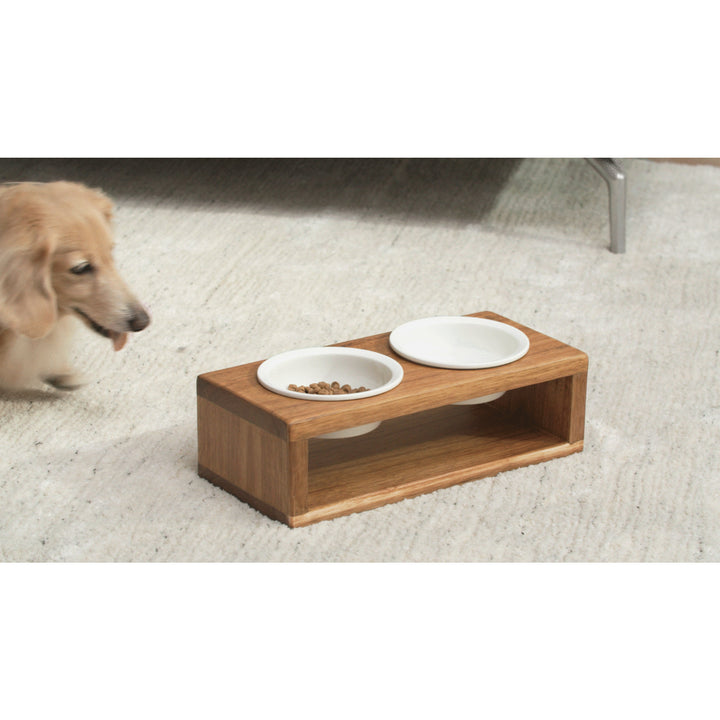Dog approaching a Teamson Pets Billie Small Elevated Wood Pet Feeder with Ceramic Bowls, Brown on a carpeted floor.