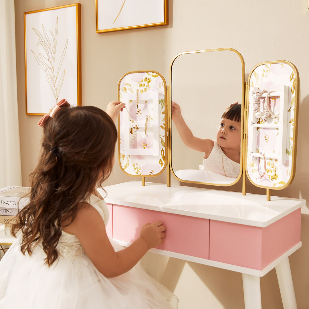 A little girl hanging a necklace on a rack next to the mirror of her pink and white vanity set.