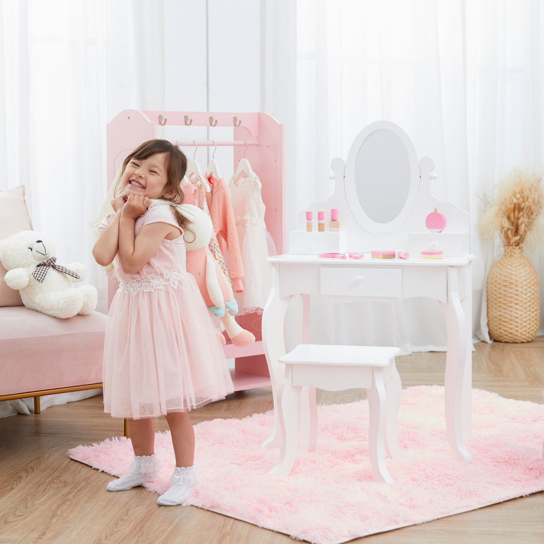 A little girl in a pink dress posing next to the white vanity set with mirror and matching stool in a white and pink bedroom.