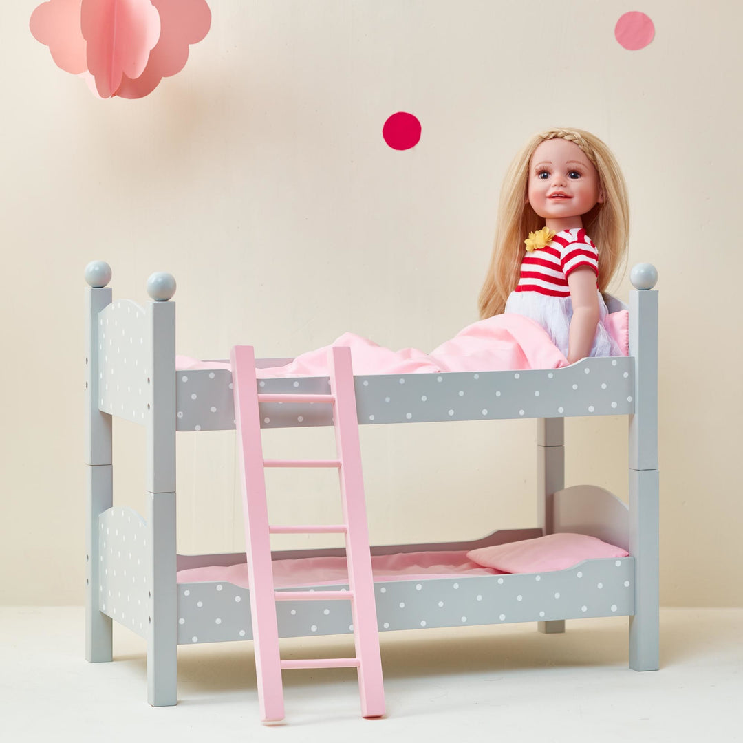 A doll in a red and white striped dress sitting on the top of the a gray with white polka dots bunk bed with pink bedding, pink ladder.