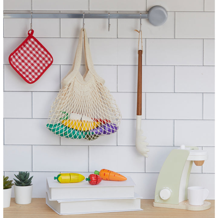 The Little Chef Frankfurt Wooden Cutting food play kitchen accessories with filet net bag is hanging on the backsplash of a kitchen counter, some veggies are displayed on the counter.