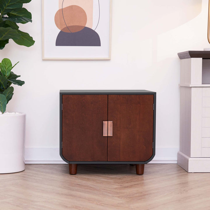 Teamson Pets Small Dyad Wooden Cat Litter Box Enclosure and Side Table, Mocha Walnut, sat up against a white wall next to a tall potted plant.
