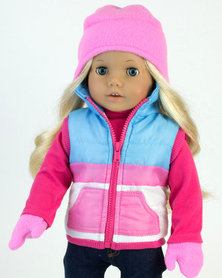 A blonde 18" doll with blue eyes dressed in a pink hat and gloves, blue and pink ski vest, blue and pink snow boots, and navy leggings