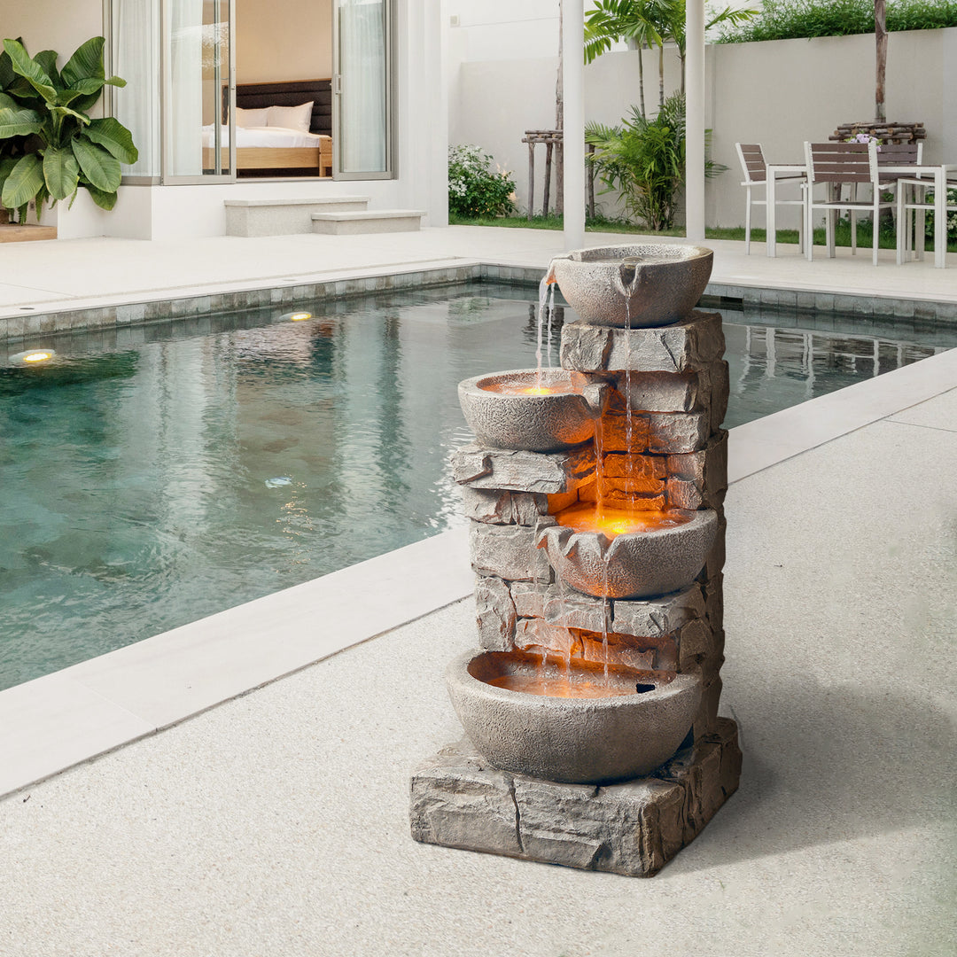 Teamson Home Outdoor Cascading Bowls & Stacked Stone water fountain with the lights on, next to a swimming pool
