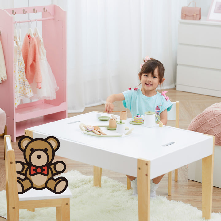 A little girl sitting at a white and wood child-sized table with an illustrated teddy bear across from her.