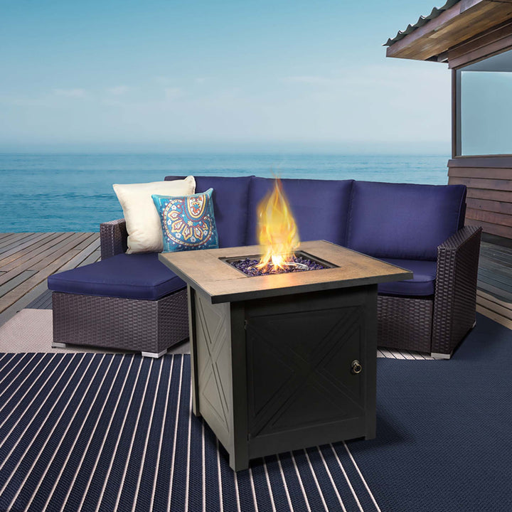 Teamson Home Outdoor Square 30" Propane Gas Fire Pit with Steel Base, Black, next to a PE wicker sofa with a seaside view behind it