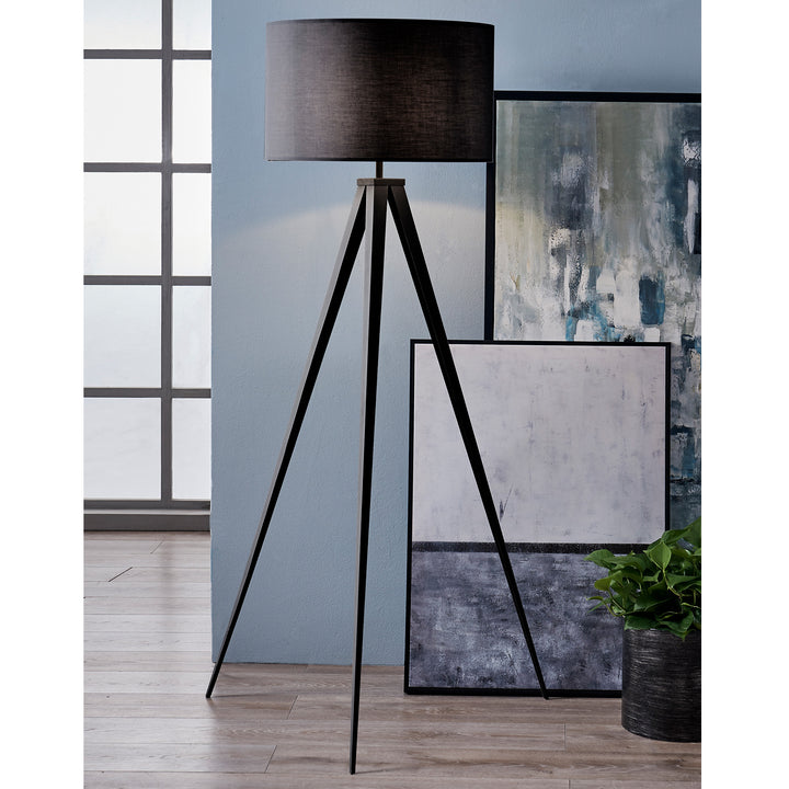 A sleek, modern Teamson Home Romanza 60" Postmodern Tripod Floor Lamp with Drum Shade, Matte Black stands next to a cluster of abstract paintings leaning against a blue wall, with a potted plant at the corner.