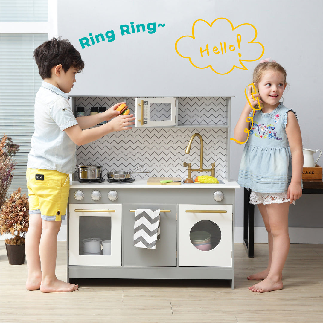 Two children enjoy their Teamson Kids Little Chef Berlin Modern Play Kitchen with 6 Accessories, Gray/White, with ample storage space for accessories, as the boy pretends to cook and the girl pretends to talk on a toy phone equipped with interactive features.