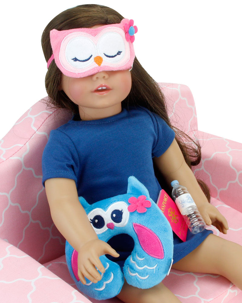 An 18" brunette doll sits on a pink sofa with an owl sleep mask over her eyes, with her travel pillow, passport, and water bottle lay in her lap.