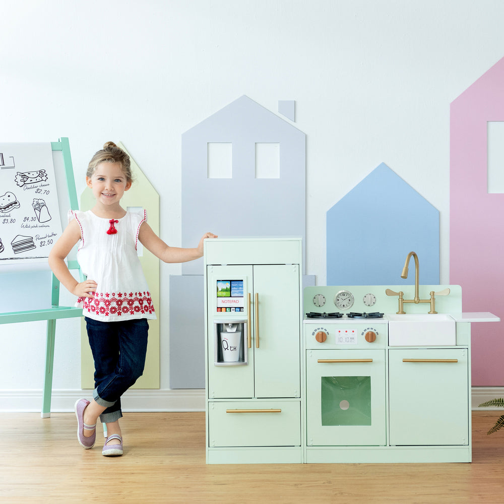 A young girl standing proudly next to a Teamson Kids Little Chef Charlotte Modern Play Kitchen in Mint/Gold in a colorful playroom.