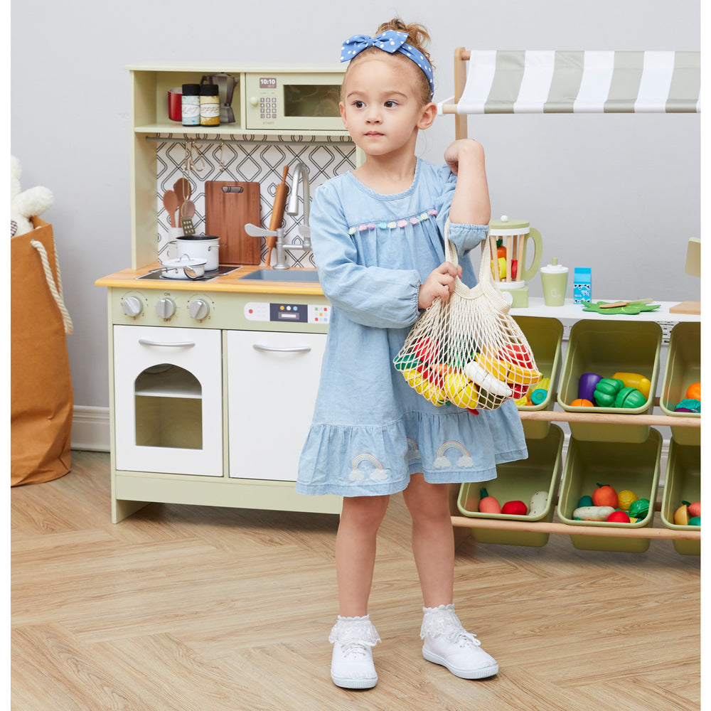 A girl stands in a playroom with her Little Chef Frankfurt Wooden Cutting food play kitchen accessories with filet net bag