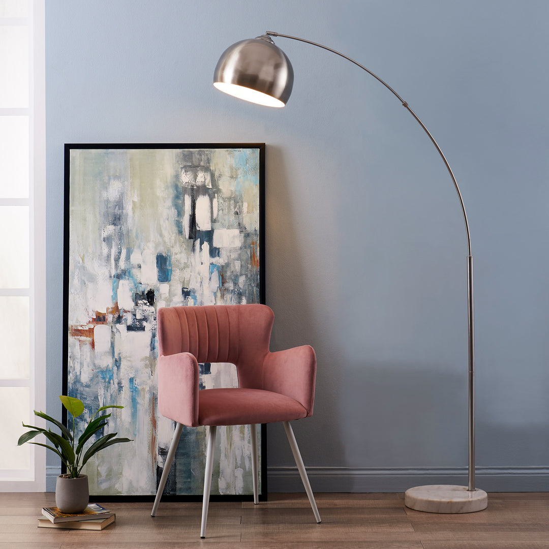A pink chair and a Teamson Home Arquer Arc 68" Metal Floor Lamp with Bell Shade, Polished Nickel, designed to illuminate a reading nook or office with soft light, in front of a painting.