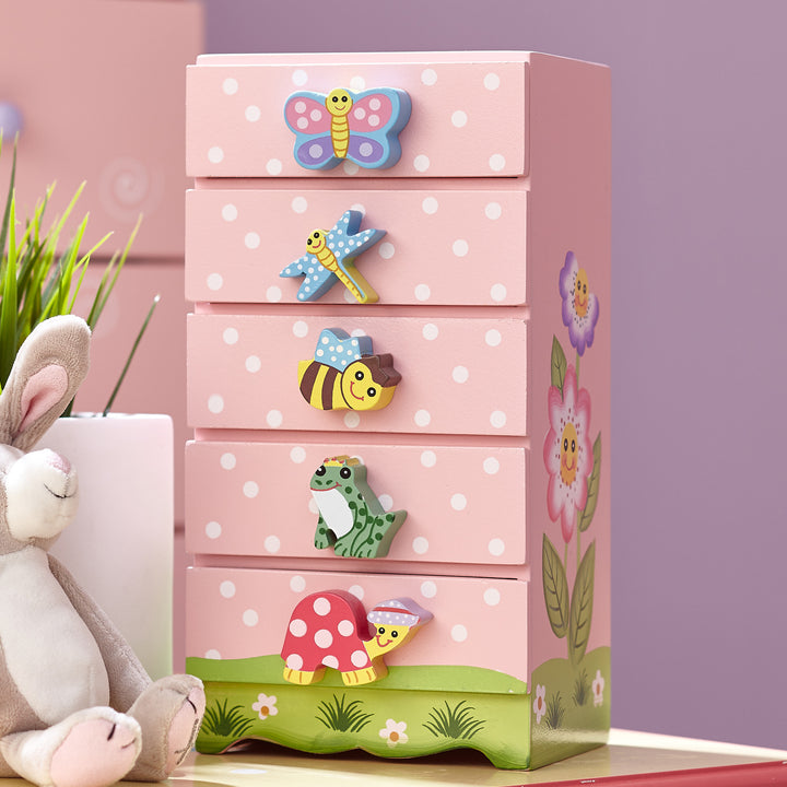 A Fantasy Fields Magic Garden Kids Wooden Trinket Chest, Pink with a bunny next to it and drawer pulls of a butterfly, dragonfly, bee, frog, and turtle on a pink and white polka dotted surface.