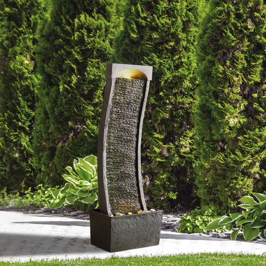 Teamson Home Indoor/Outdoor Modern Curved Slate Waterfall Fountain with LED Lights set outside against shrubs in a landscaped area