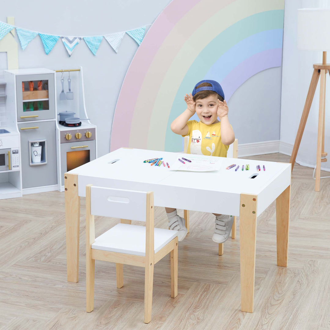 A little boy in a playroom sitting at a white and wood child-sized table and two chairs. 
