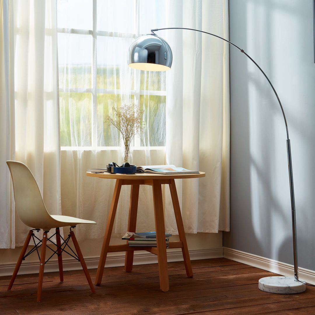 A table with a Teamson Home Arquer Arc 66" Metal Floor Lamp with Bell Shade, Chrome and a chair in front of a window.