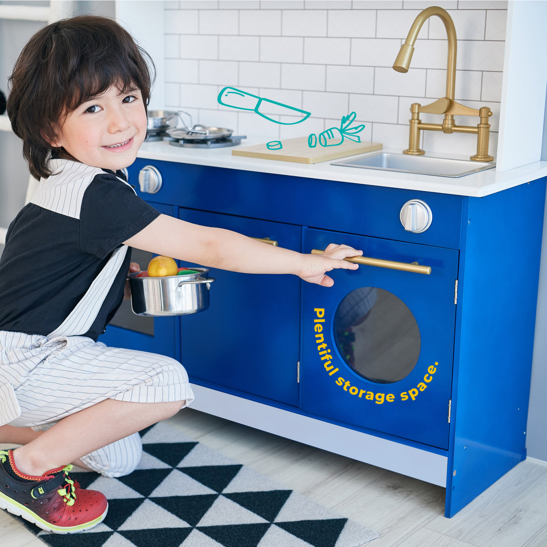 Child posing with a Teamson Kids Little Chef Berlin Play Kitchen with Cookware Accessories, White/Blue, showcasing playful storage space.
