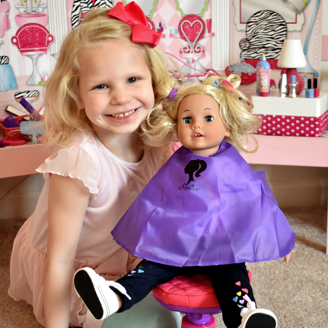 A little girl smiling next to her 18" blonde doll who is sitting in a salon chair with a purple smock on.