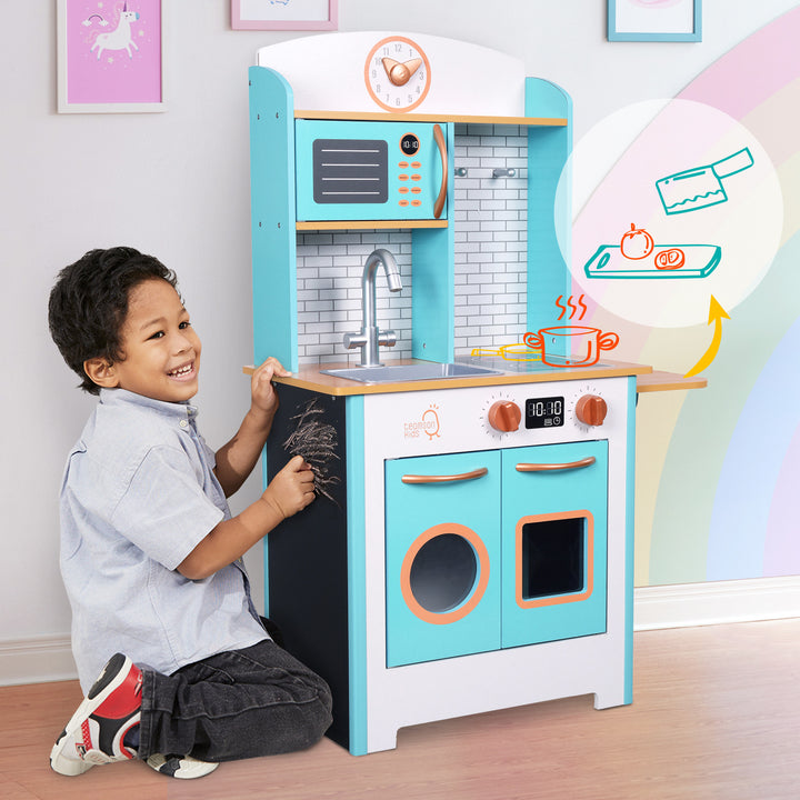 A young boy writing on the chalkboard side panel of the  Teamson Kids Little Chef Santos Retro Wooden Kitchen Playset, Aqua/White.