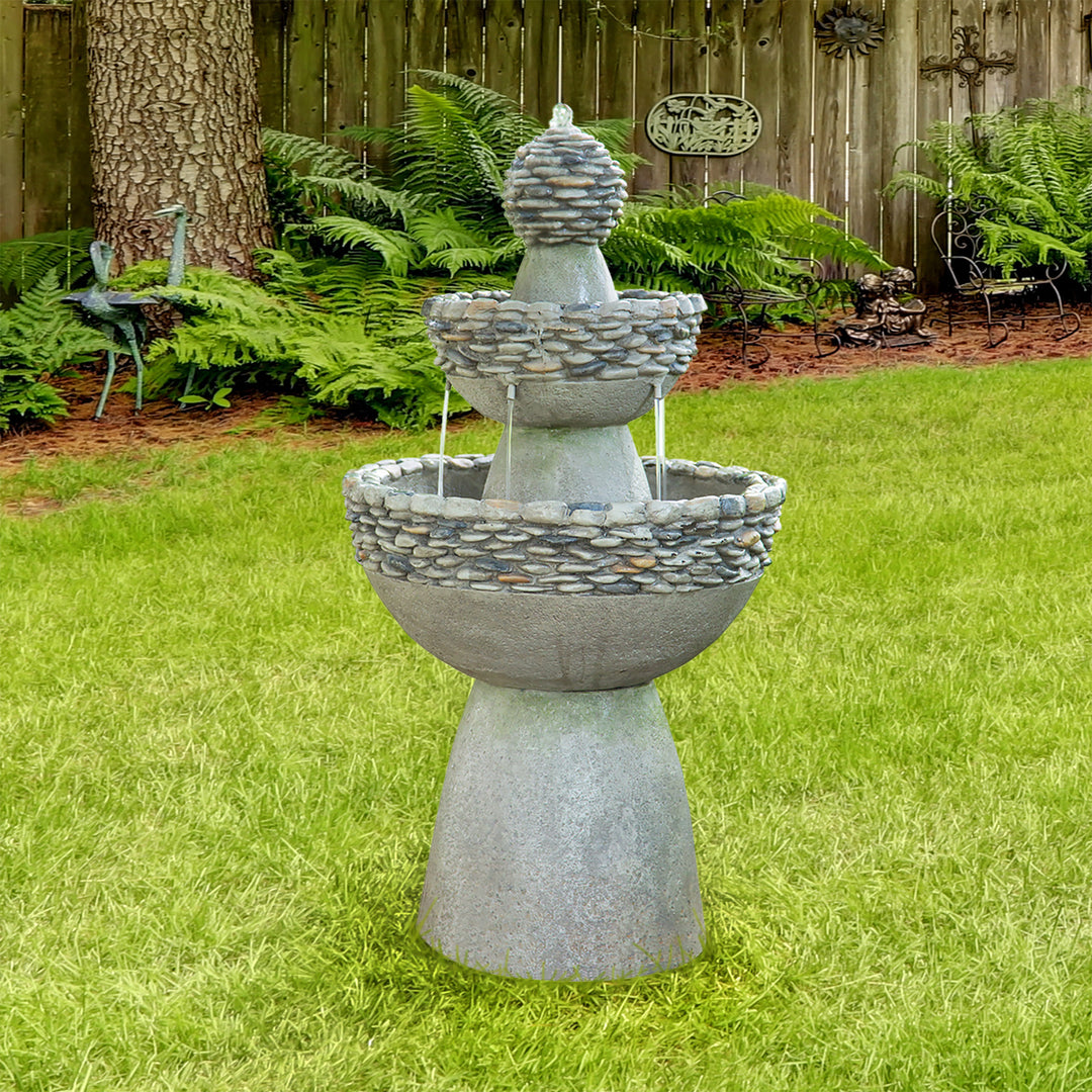 A Teamson Home Outdoor 3-Tier Pedestal Floor Fountain, Gray, standing outside in a backyard garden with ferns and a wooden fence behind it