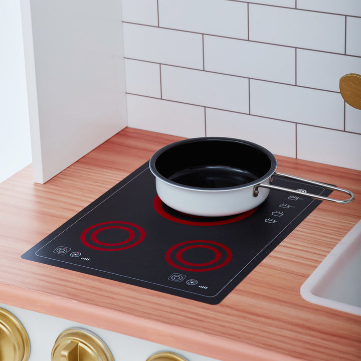A black frying pan on an induction stove top in a modern kitchen designed as a Teamson Kids Little Chef Boston Classic Play Kitchen & Cookware, White.
