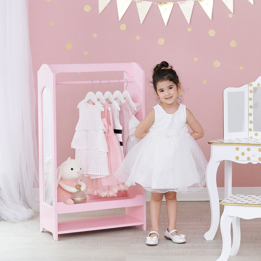 A little girl dressed in a white dress standing between a pink wardrobe with dresses hanging from it and a stuffed bunny and a white vanity table and stool with gold polka dots.