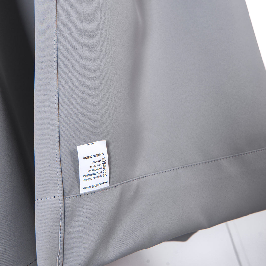 Close-up of a 96" Lamia Premium Solid Insulated Thermal Blackout Grommet Window Panels, Fossil Grey label on a grey polyester fabric showing care instructions and fabric composition.