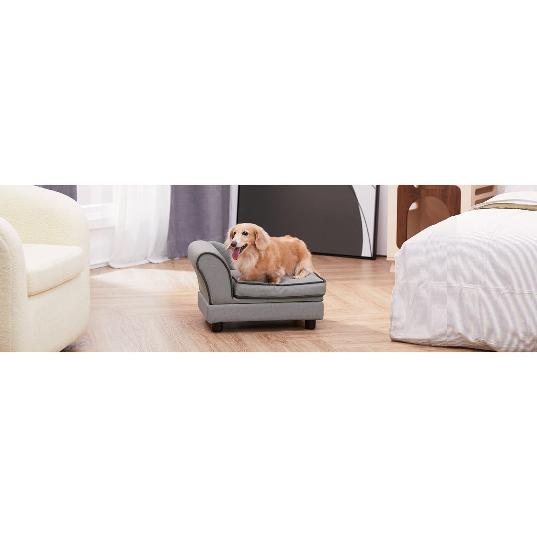 A small dog sitting on a small gray chaise lounge pet bed placed inside a bedroom.