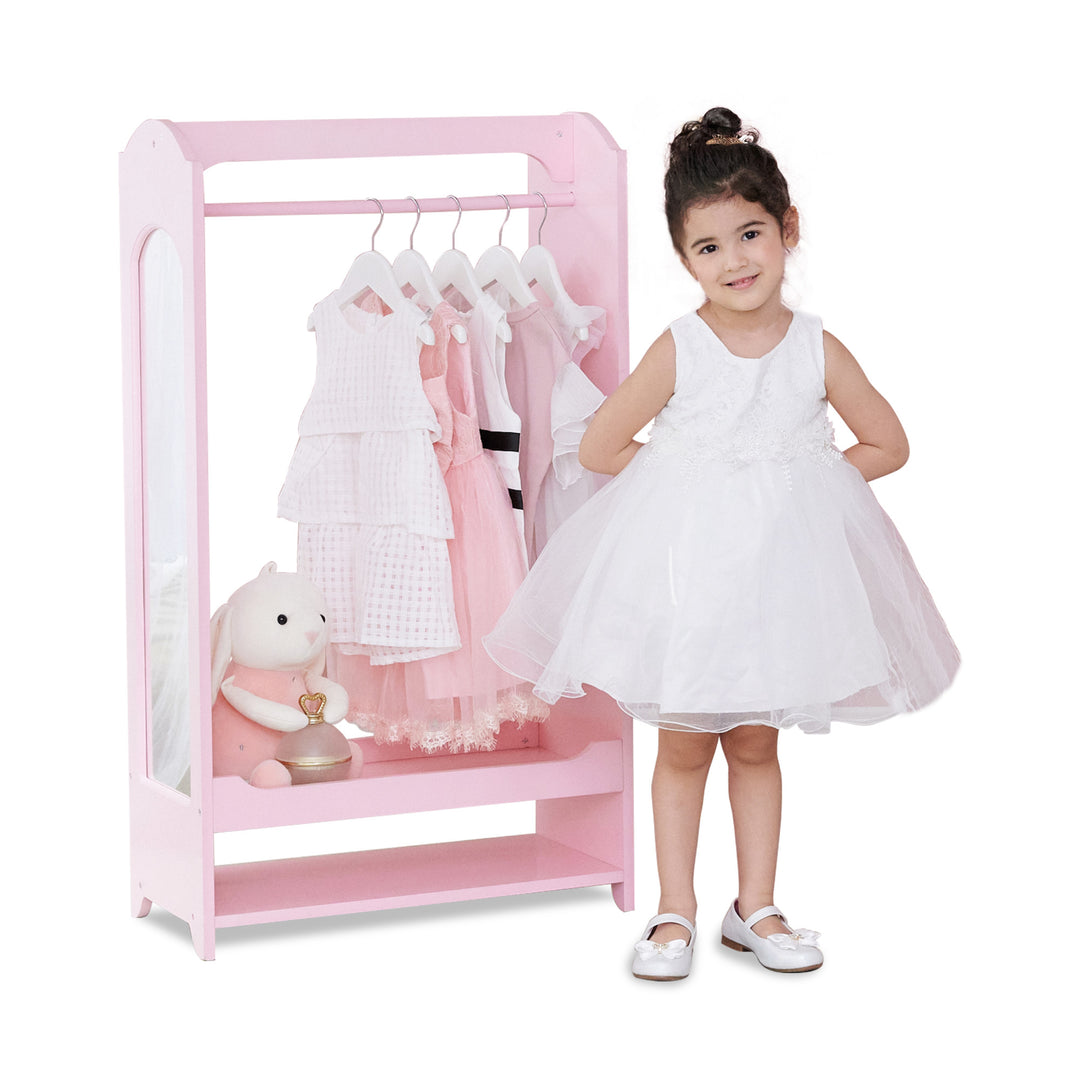 A little girl dressed in a white dress standing in front of a pink wardrobe with dresses hanging from it.