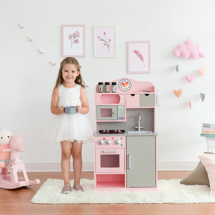 A young girl smiling in a playroom with a Teamson Kids Little Chef Florence Classic Play Kitchen, Pink/Gray and wall decorations.