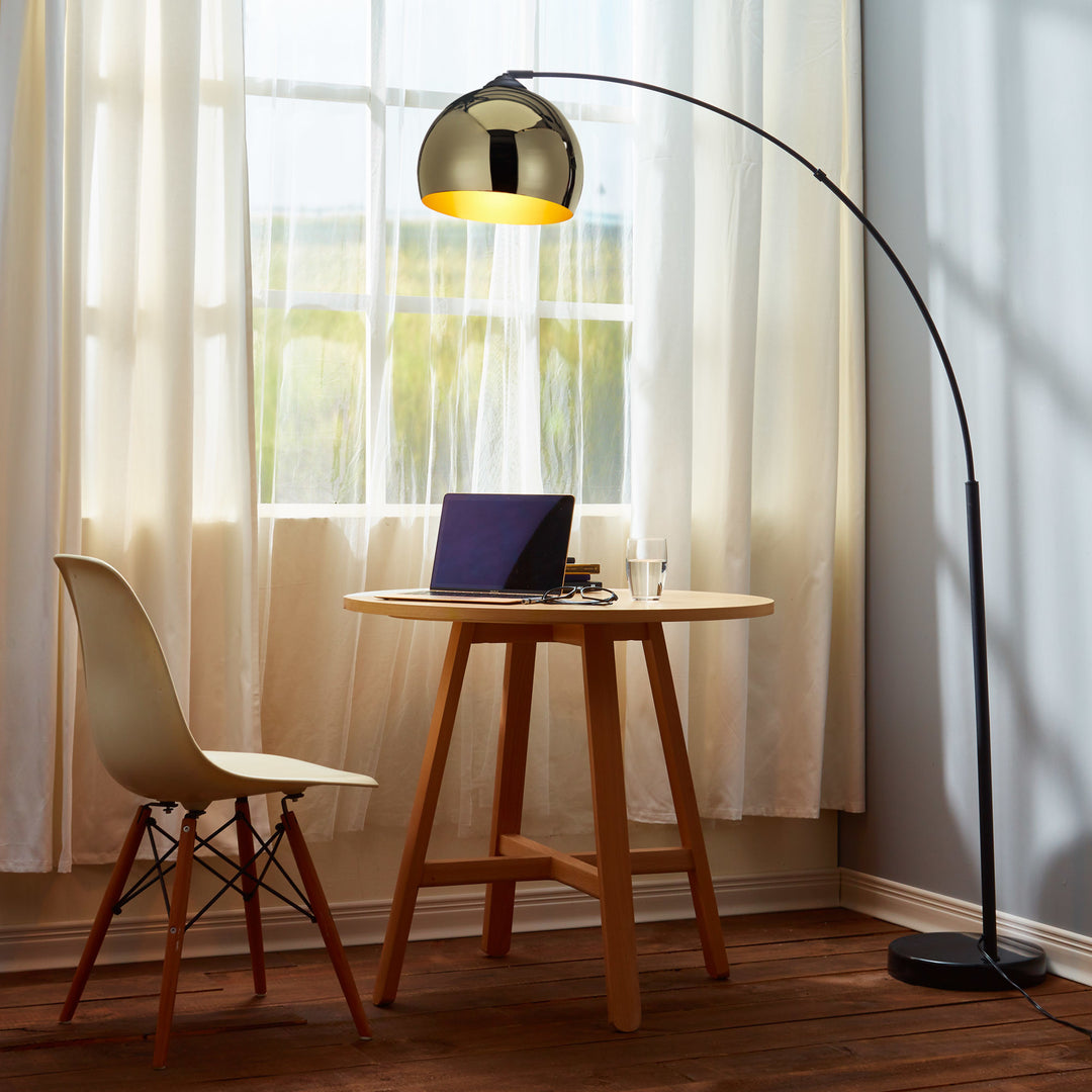 A table with a Teamson Home Arquer Arc Metal Floor Lamp with Bell Shade, Gold, having a marble base and bell shade, and a chair in front of a window.